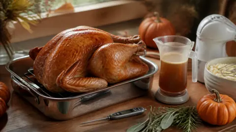 A golden roast turkey on a rack in a stainless steel roasting pan on a wooden counter with a digital meat thermometer, a gravy separator, a hand mixer, mashed potatoes and fresh herbs
